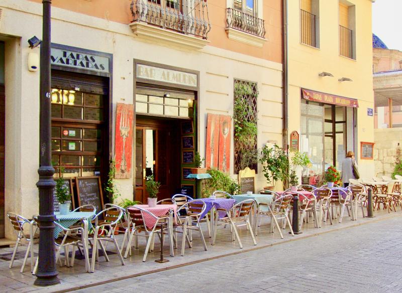 Exterior view of a traditional Spanish tapas bar with outdoor terrace seating, checkered tablecloths, and chalkboard menus.