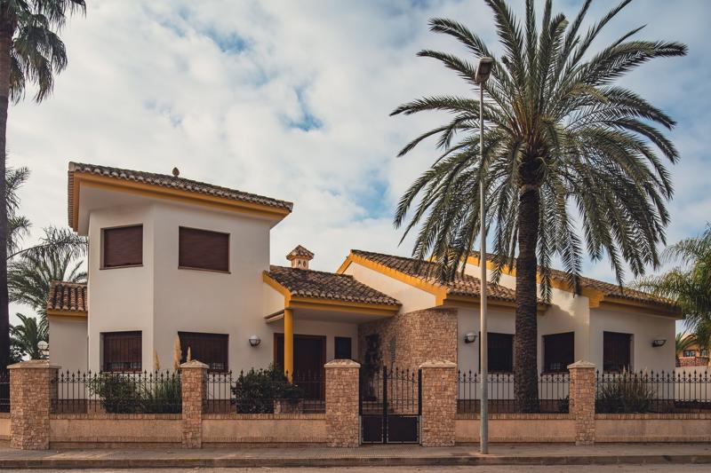 A modern Spanish detached villa with white walls, a traditional tiled roof, a prominent palm tree, and a stone-and-iron perimeter fence, representing premium family real estate in a prime school catchment zone.