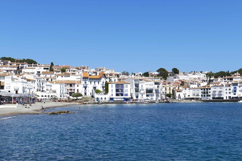 The iconic waterfront of Cadaqués, displaying a row of traditional whitewashed buildings with blue accents situated directly on the water's edge under a clear blue sky.
