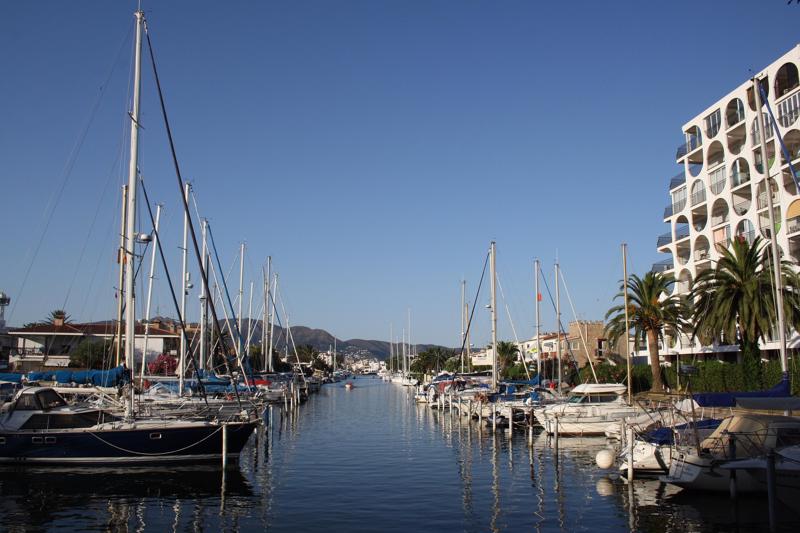 A wide residential canal in Empuriabrava lined with moored sailboats and motorboats, framed by palm trees and modern white apartment buildings.