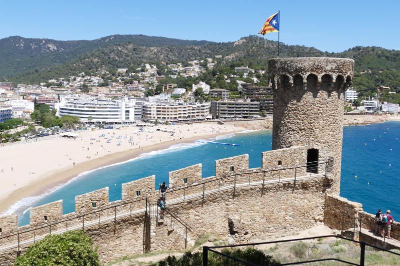A view from the ancient stone battlements and fortress tower of Tossa de Mar, looking down onto a sweeping sandy beach, a bright blue Mediterranean sea, and coastal apartments.