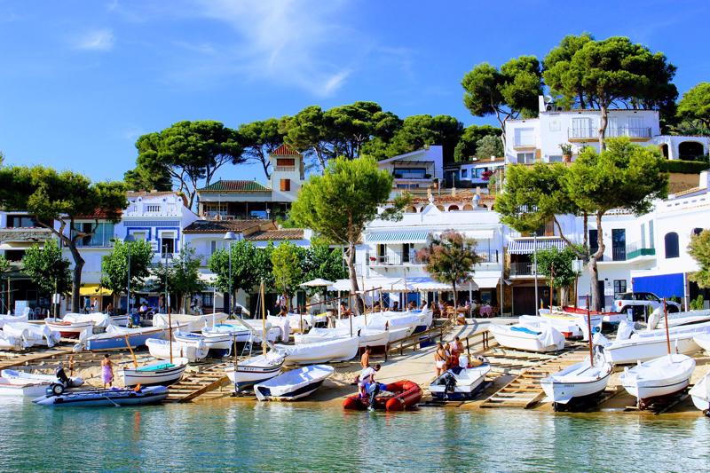 A picturesque Costa Brava coastal village featuring traditional white houses, pine trees, and small boats resting on wooden ramps by the water.