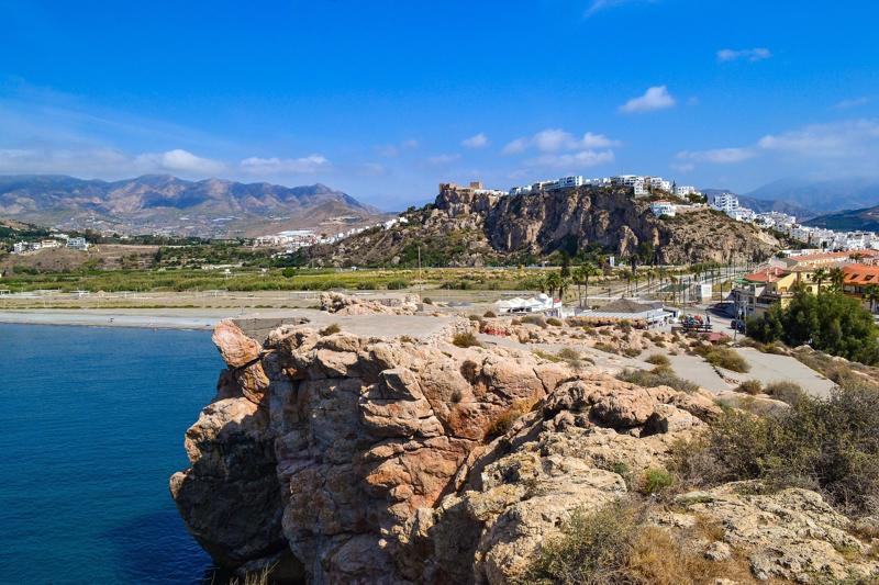 A panoramic view from a rocky coastal cliff featuring a Moorish castle on a hilltop, the blue Mediterranean sea, and the Sierra Nevada mountain foothills in the background.