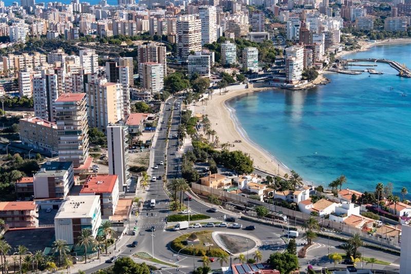 An aerial view of a densely populated Spanish coastal city featuring high-rise apartment buildings along a curved sandy beach, adjacent to a main coastal road and roundabout next to the bright blue Mediterranean Sea.