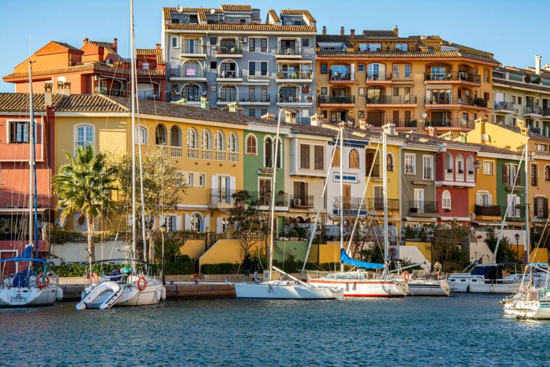 A picturesque Spanish marina lined with vibrant, multi-colored waterfront townhouses and apartments, with several white sailboats docked in the calm blue water under a clear sunny sky.