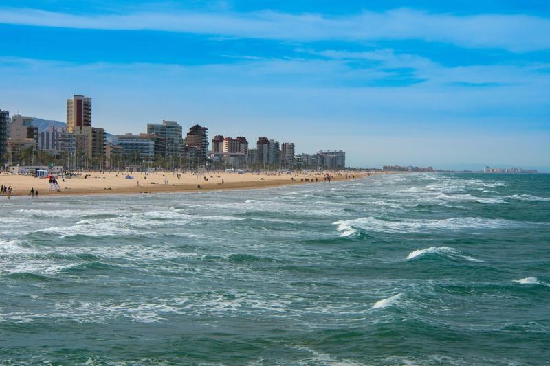 Wide sandy beach in Valencia province lined with seafront apartment buildings and gentle Mediterranean waves under a clear blue sky.