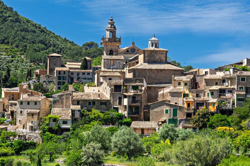 A historic Mallorcan stone village nestled among lush green hills in the Tramuntana mountains, centred around a traditional church with a tall bell tower.