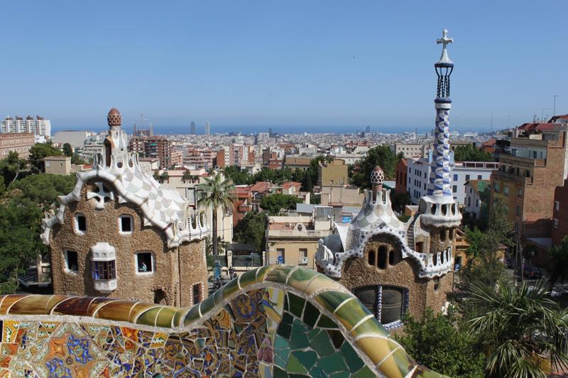 Panoramic view of Barcelona from Park Güell's serpentine mosaic bench, overlooking Antoni Gaudí's modernist pavilion rooftops, the dense city centre, and the Mediterranean Sea under a clear blue sky.