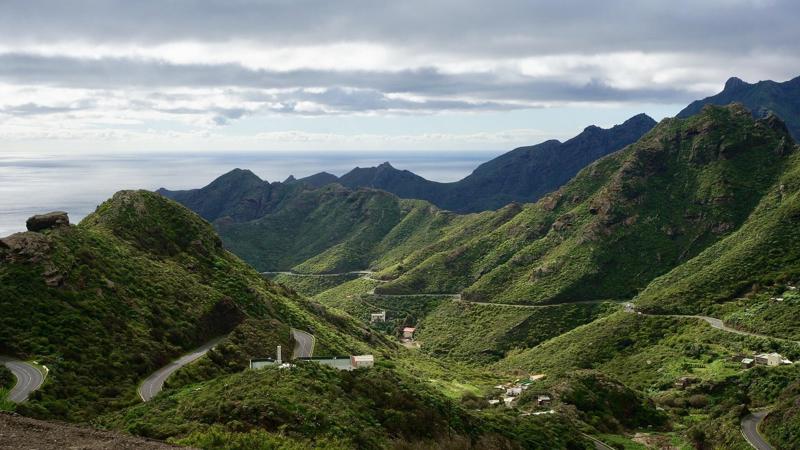 A paved road winds through the lush green volcanic mountains of the Anaga Rural Park in north Tenerife under a cloudy sky.